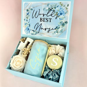 May include: A light blue gift box with the text "World's Best Nurse" and floral accents. Inside, a glass tumbler with a name, a pen, a compact mirror, a rose, and other accessories are visible.