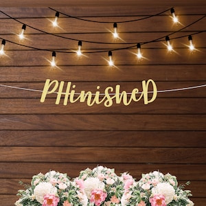 May include: Gold glitter banner with the word "PHInished" hanging on a wood wall with string lights. The banner is hanging above a bouquet of white and pink flowers.