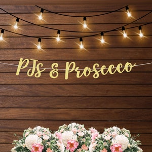 May include: Gold glitter banner with the text "PJs & Prosecco" hanging on a wood wall with string lights.  There are three bouquets of white and pink flowers in the foreground.
