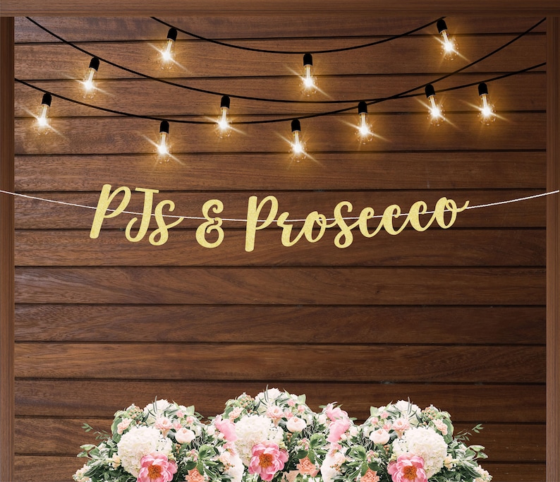 May include: Gold glitter banner with the text "PJs & Prosecco" hanging on a wood wall with string lights.  There are three bouquets of white and pink flowers in the foreground.