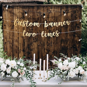 May include: A rustic wedding table setting with white flowers, greenery, and candles. A gold banner with the text "Custom banner two lines" hangs on a wooden wall behind the table.