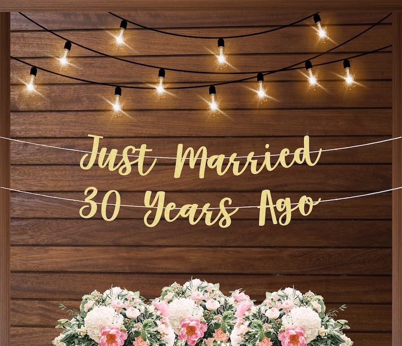 May include: Gold glitter banner with the text "Just Married 30 Years Ago" hanging on a wood wall with string lights.