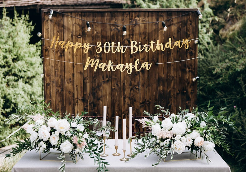 K&ouml;nnte beinhalten: Ein rustikaler Holz-Hintergrund mit einem goldenen Glitzerbanner mit der Aufschrift "Happy 30th Birthday Makayla". Zwei wei&szlig;e Blumenarrangements mit Gr&uuml;n sind auf einem Tisch vor dem Hintergrund.