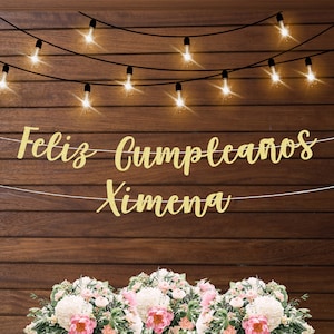 Könnte beinhalten: Goldene Glitzergirlande mit dem Text "Feliz Cumpleanos Ximena" an einer Holztafel mit Lichterketten. Die Girlande ist mit Blumen verziert.