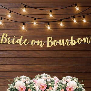 May include: Gold glitter banner with the text "Bride on Bourbon" hanging on a wood wall with string lights.