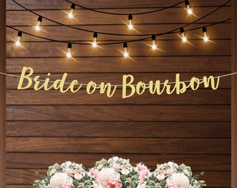 May include: Gold glitter banner with the text "Bride on Bourbon" hanging on a wood wall with string lights.