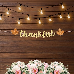 May include: A gold glitter banner with the word "thankful" and two orange glitter maple leaves hanging on a brown wood wall with a string of white lights.