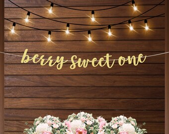 May include: A gold glitter banner that reads "berry sweet one" hangs in front of a wooden background. String lights are strung above the banner. A floral arrangement with pink and white flowers is at the bottom.