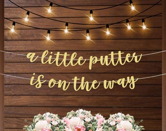 Könnte beinhalten: Ein goldener Glitzerbanner mit dem Text "a little putter is on the way" hängt an einer Holzwand mit Lichterketten. Der Banner ist mit Blumen verziert.