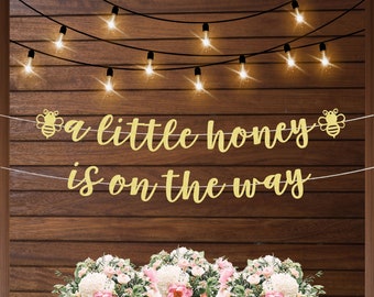 Puede incluir: Pancarta dorada con purpurina con el texto "a little honey is on the way" y dos ilustraciones de abejas. La pancarta está colgada en un fondo de madera con luces de cadena.