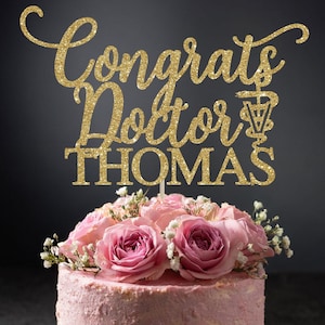 May include: Gold glitter cake topper with the text "Congrats Doctor THOMAS" and a caduceus symbol. The topper is on a pink frosted cake with pink roses.