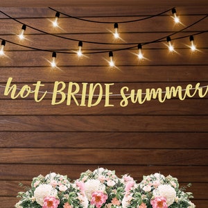 K&ouml;nnte beinhalten: Goldene Glitzergirlande mit dem Text "hot BRIDE summer" an einer Holzwand mit Lichterketten. Die Girlande ist mit Blumen verziert.