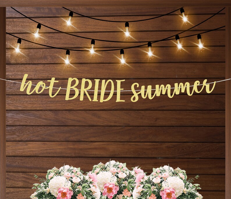 K&ouml;nnte beinhalten: Goldene Glitzergirlande mit dem Text "hot BRIDE summer" an einer Holzwand mit Lichterketten. Die Girlande ist mit Blumen verziert.