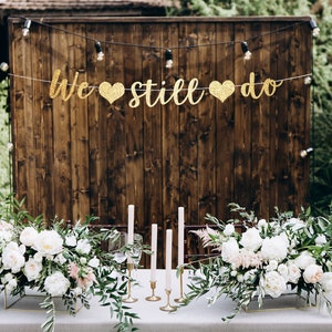 May include: A rustic wedding reception table setting with white flowers, greenery, candles, and a gold glitter banner that reads "We still do".