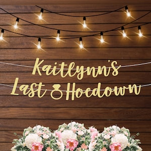 以下が含まれることがあります： ダイヤモンドの指輪のシンボルが付いた「Kaitlynn's Last Hoedown」という文字が書かれた金色のグリッターバナー。バナーは、ストリングライトがついた木製の壁にかけられています。