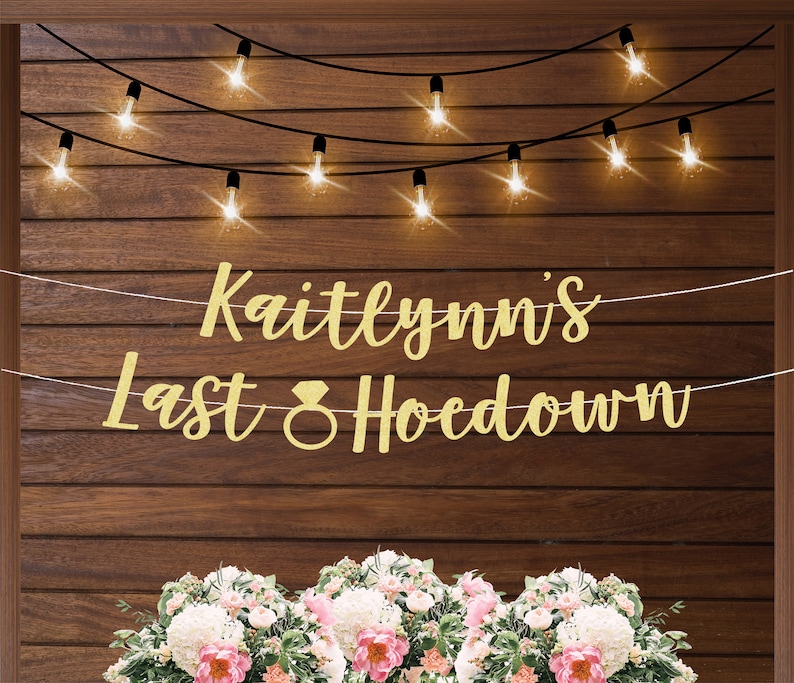 以下が含まれることがあります： ダイヤモンドの指輪のシンボルが付いた「Kaitlynn's Last Hoedown」という文字が書かれた金色のグリッターバナー。バナーは、ストリングライトがついた木製の壁にかけられています。