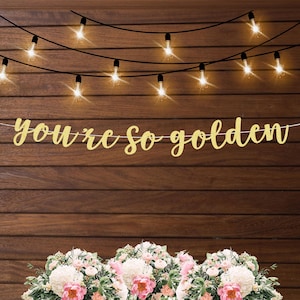 May include: Gold glitter banner with the text "You're so golden" hanging on a wood wall with string lights.