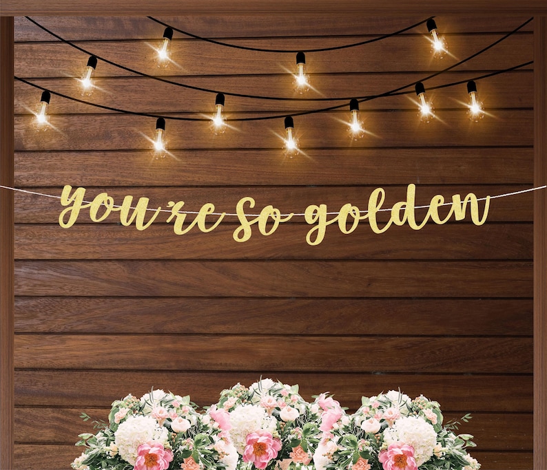 May include: Gold glitter banner with the text "You're so golden" hanging on a wood wall with string lights.