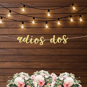 May include: Gold glitter "adios dos" banner hanging from a string of lights against a wood background.  Two bouquets of white and pink flowers are in the foreground.