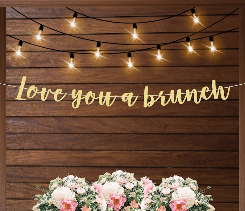 May include: Gold glitter banner with the text "Love you a brunch" hanging on a wood wall with string lights.
