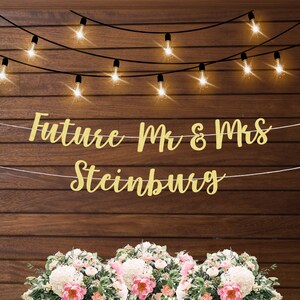 May include: Gold glitter banner with the text "Future Mr & Mrs Steinberg" hanging on a wood wall with string lights. The banner is hanging above a bouquet of white and pink flowers.