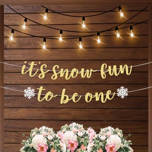 May include: A festive banner with the words "it's snow fun to be one" in gold script, adorned with snowflake accents. The banner hangs against a wooden backdrop, complemented by string lights and a floral arrangement.