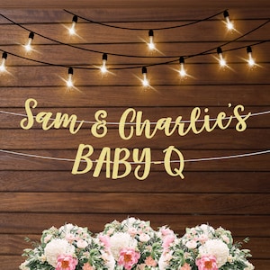 Puede incluir: Una pancarta dorada con purpurina con el texto "Sam & Charlie's BABY-Q" colgada en una pared de madera con luces de cadena. La pancarta est&aacute; decorada con flores blancas.