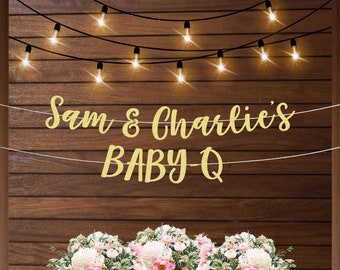 May include: A gold glitter banner with the text "Sam & Charlie's BABY-Q" hanging on a wood wall with string lights. The banner is decorated with white flowers.