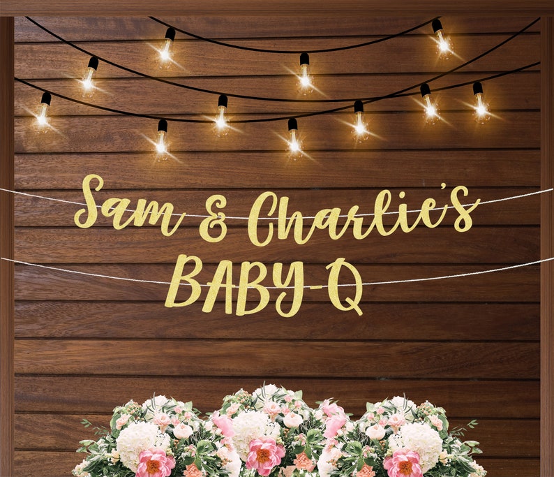 Puede incluir: Una pancarta dorada con purpurina con el texto "Sam & Charlie's BABY-Q" colgada en una pared de madera con luces de cadena. La pancarta est&aacute; decorada con flores blancas.