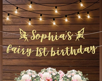 May include: Gold glitter banner with the text "Sophia's fairy 1st birthday" and two fairy silhouettes. The banner is hanging on a wood wall with string lights.