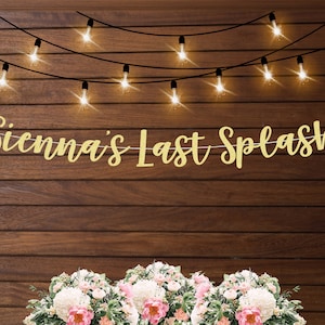 May include: Gold glitter banner with the text "Sienna's Last Splash" hanging on a wooden wall with fairy lights. The banner is decorated with flowers.