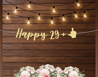 May include: Gold glitter banner with the text "Happy 29+" and a middle finger emoji. The banner is hanging on a wood wall with string lights.