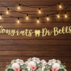 May include: Gold glitter banner with the text "Congrats Dr Bells" and a tooth icon. The banner is hanging from a string of lights against a wood background.