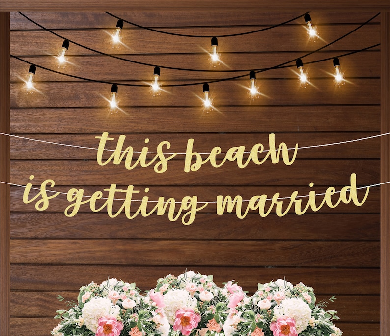 Może przedstawiać: Złota girlanda z błyskotkami z napisem "this beach is getting married" wisząca na drewnianej ścianie z lampkami choinkowymi.
