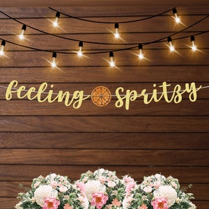 Puede incluir: Una pancarta dorada con la frase "feeling spritzy" y un gr&aacute;fico de una rodaja de c&iacute;trico. Est&aacute; colgada sobre un fondo de madera, con luces de cadena arriba y un arreglo floral abajo.