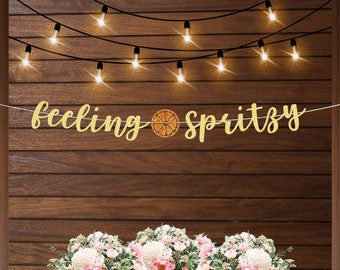 May include: A banner with the words "feeling spritzy" in gold script, with a citrus slice graphic. The banner is strung across a wooden background, with string lights above and a floral arrangement below.