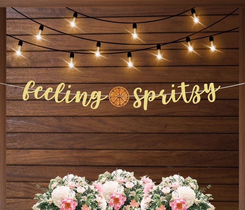 Puede incluir: Una pancarta dorada con la frase "feeling spritzy" y un gr&aacute;fico de una rodaja de c&iacute;trico. Est&aacute; colgada sobre un fondo de madera, con luces de cadena arriba y un arreglo floral abajo.