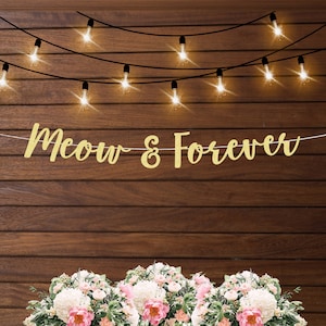 May include: A gold glitter banner with the words "Meow & Forever" strung across a wooden background. String lights are above the banner. A floral arrangement with pink and white flowers is at the bottom.