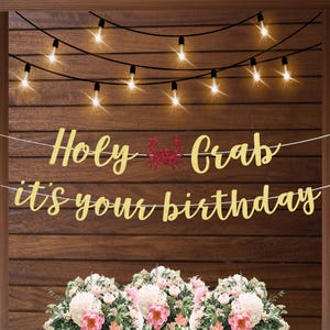 May include: A gold glitter banner with the text Holy Crab it's your birthday, featuring a red glitter crab icon between the words Holy and Crab, suspended on white string against a dark wood background with glowing string lights above.