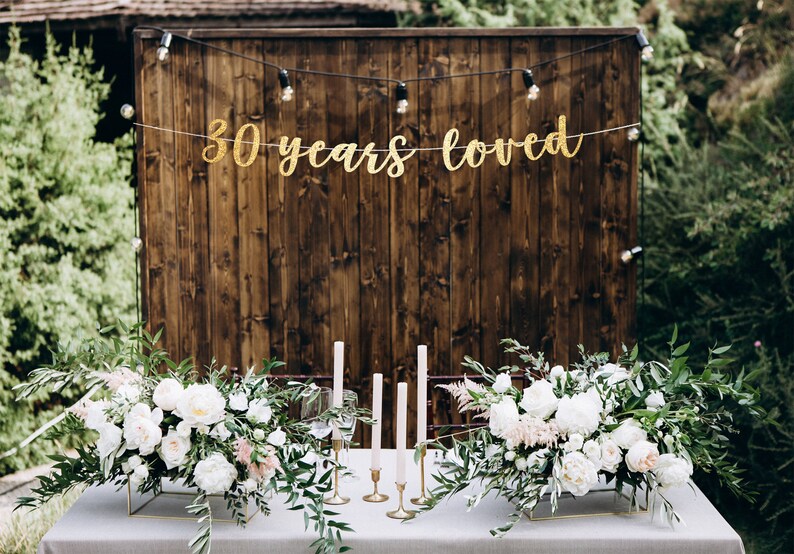 May include: A wedding table setting with white flowers and greenery in gold square vases. There are candles on the table and a banner that says "30 years loved" hanging on a wooden wall behind the table.