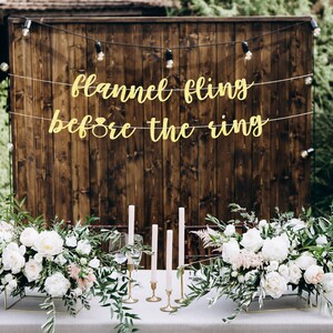 May include: A rustic wooden backdrop with a gold banner that reads "Flannel Fling Before the Ring". Two centerpieces with white flowers and greenery are on a table in front of the backdrop.