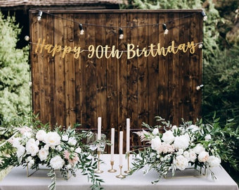 May include: A rustic wooden backdrop with a gold glitter banner that reads "Happy 90th Birthday". Two tables are set with white flowers, greenery, and candles.