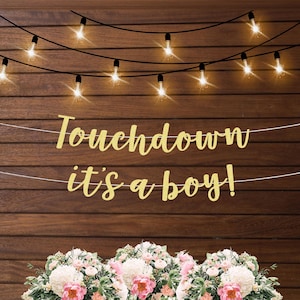 K&ouml;nnte beinhalten: Goldene Glitzergirlande mit dem Text "Touchdown es ist ein Junge!" an einer Holztafel mit Lichterketten.