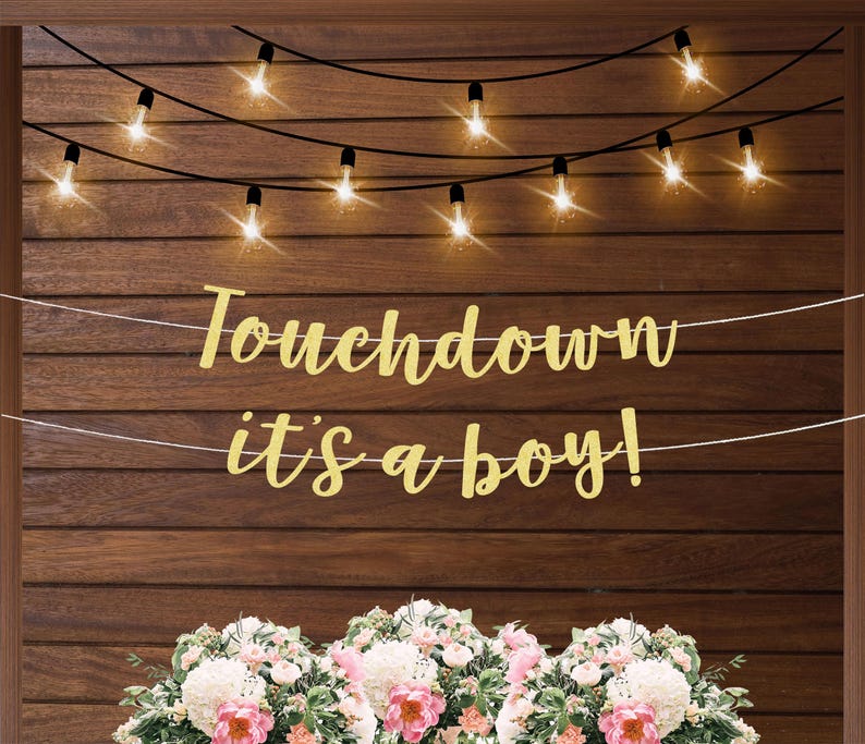 K&ouml;nnte beinhalten: Goldene Glitzergirlande mit dem Text "Touchdown es ist ein Junge!" an einer Holztafel mit Lichterketten.