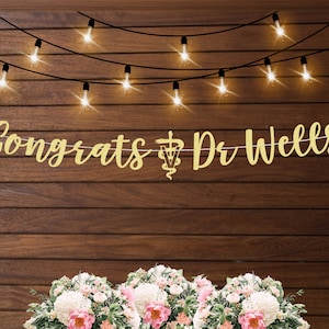 May include: Gold glitter banner with the text "Congrats Dr. Wells" and a veterinarian symbol. The banner is hanging on a wood wall with string lights.