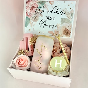 May include: A pink gift box with the words "World's Best Nurse" on the lid. Inside are a pink hair clip, a frosted glass, a gold pen, a compact mirror, a pink rose, and scrunchies.