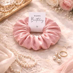 May include: A pink satin scrunchie with a white card that reads "Happy Birthday" and a small heart. The scrunchie is surrounded by pearls, roses, and jewelry, creating a celebratory theme.