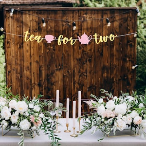 May include: A rustic wooden backdrop with a banner that reads "tea for two" decorated with pink teacups and a teapot. Two white floral arrangements with greenery are on a table in front of the backdrop. There are five gold candle holders with white candles.