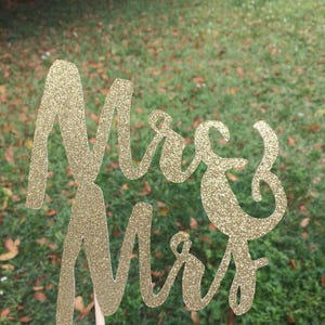 Puede incluir: Topper de pastel de brillo dorado con las palabras "Mrs & Mr" en una fuente cursiva.