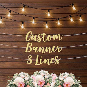 May include: Gold glitter banner with the text "Custom Banner 3 Lines" hanging on a wood background with string lights. Flowers are in the foreground.